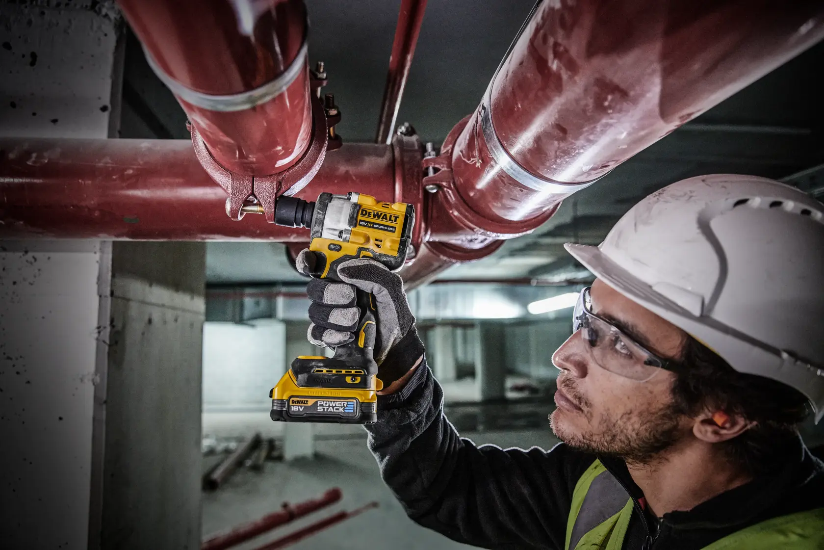 Una persona con casco de seguridad y guantes usa una llave de impacto inalámbrica DEWALT (modelo DCF921E2T) para apretar un perno en una tubería roja grande en un entorno industrial. El rostro de la persona está difuminado por privacidad.