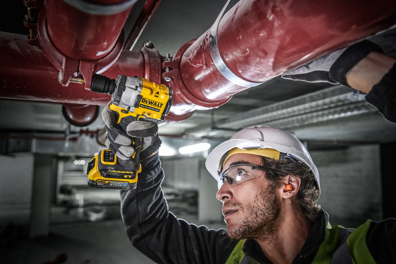 Una persona con equipo de seguridad utiliza una llave de impacto inalámbrica DEWALT (modelo DCF921E2T) para trabajar en grandes tuberías industriales rojas en un entorno interior. El rostro de la persona está difuminado.