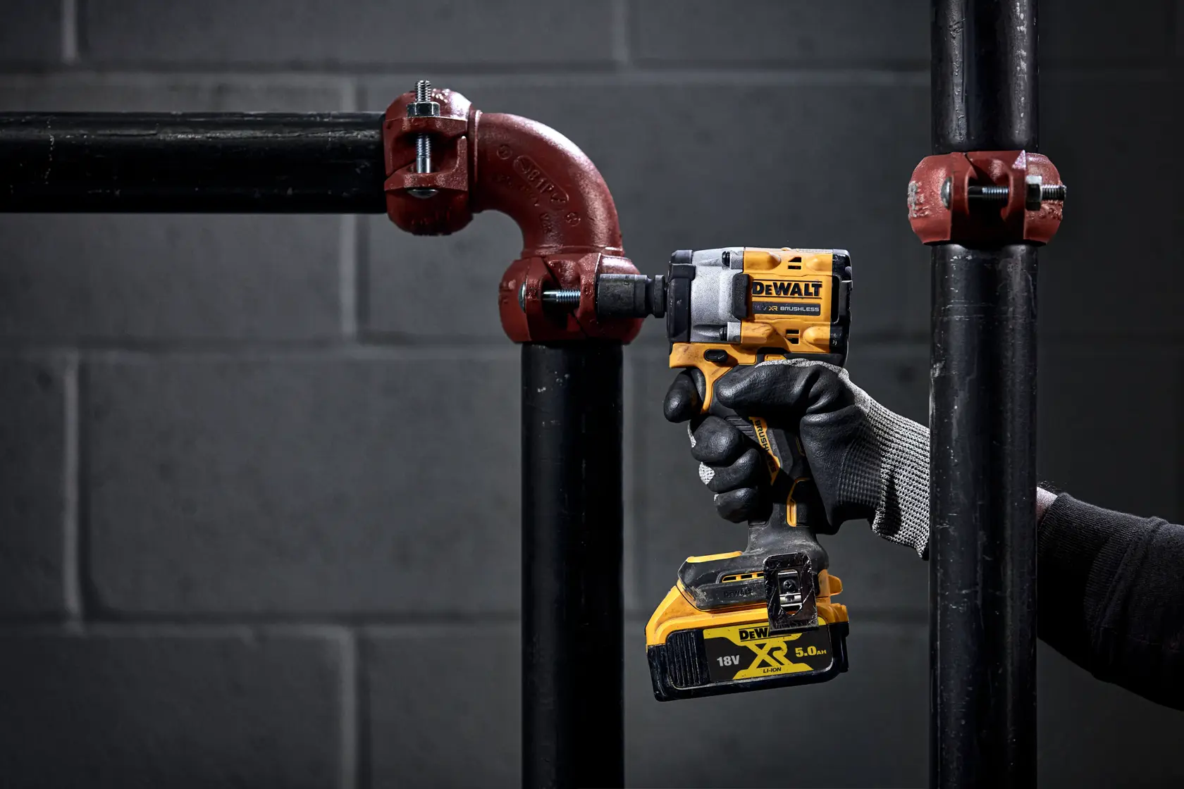A person wearing black gloves is using a yellow DEWALT DCF923N cordless impact wrench to tighten a pipe fitting. The tool is labeled with 18V and XR 5.0 Ah, and is being used on black pipes with red connectors in an industrial setting.