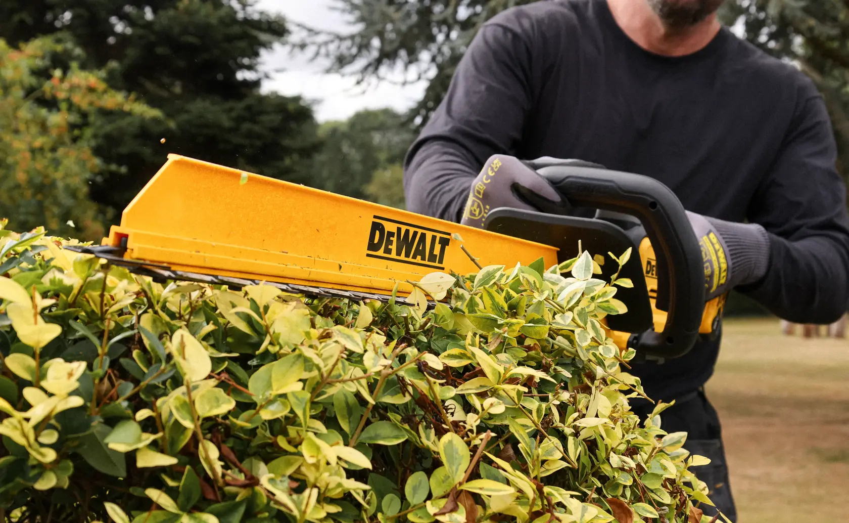 DEWALT DCMHT567N 18V hedge trimmer shown with blade sheath and sweeper attachment on a black grid background.
