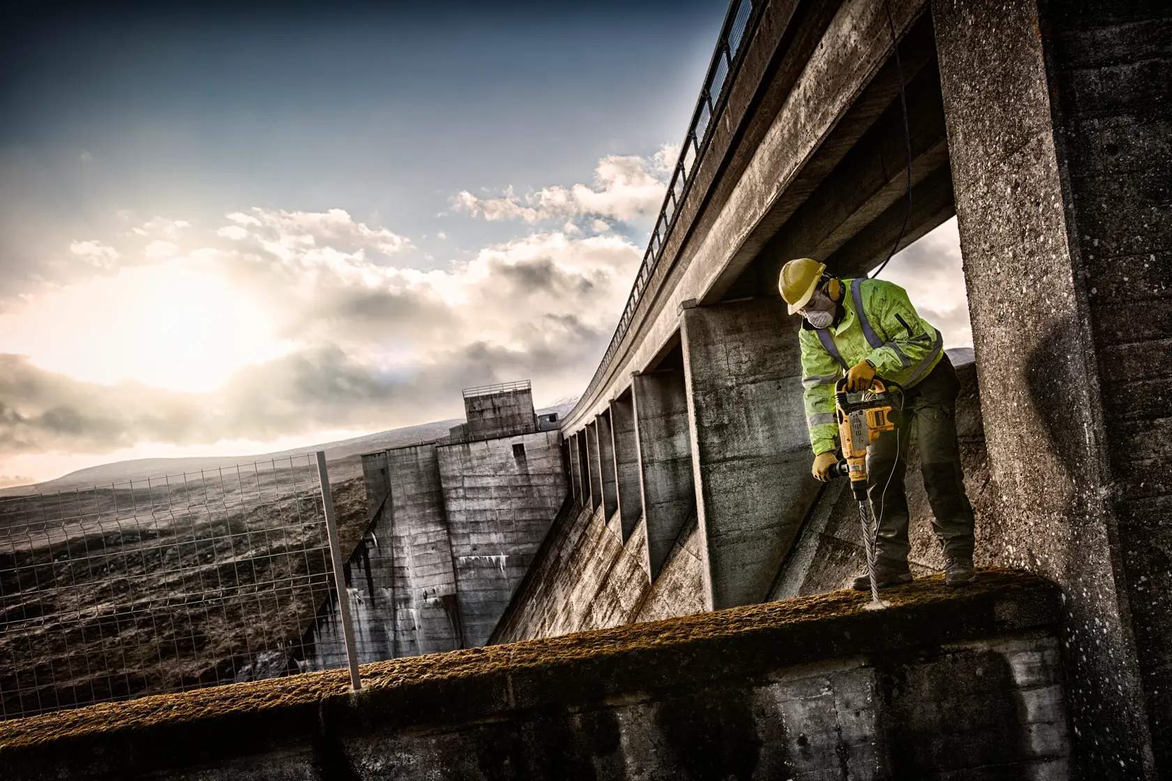 Trabajador usa un taladro DEWALT en una estructura de hormigón cerca de una presa al atardecer.