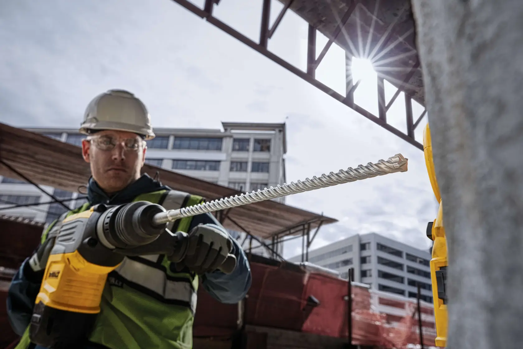 Trabajador usa taladro DEWALT con broca DT60825 en obra, con edificios de fondo.
