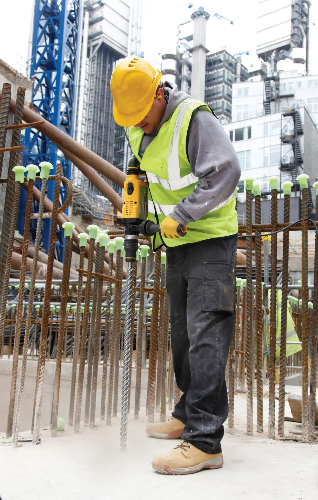Trabajador con taladro DEWALT en obra. Lleva equipo de seguridad, rodeado de varillas y edificios.