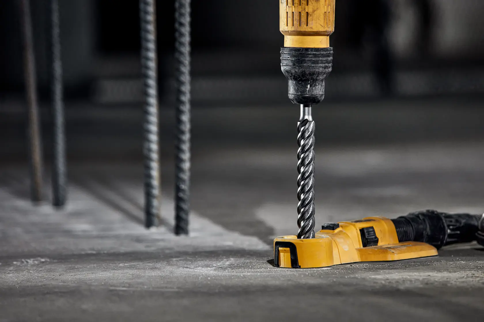Un taladro amarillo DEWALT con una broca en espiral se está utilizando para perforar una superficie de hormigón. Se ven varillas metálicas en el fondo, indicando un entorno de construcción.