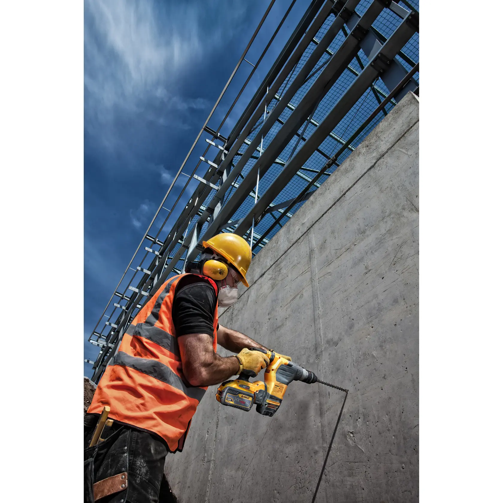 Trabajador usa taladro DEWALT en muro de concreto, lleva chaleco naranja y equipo de seguridad.