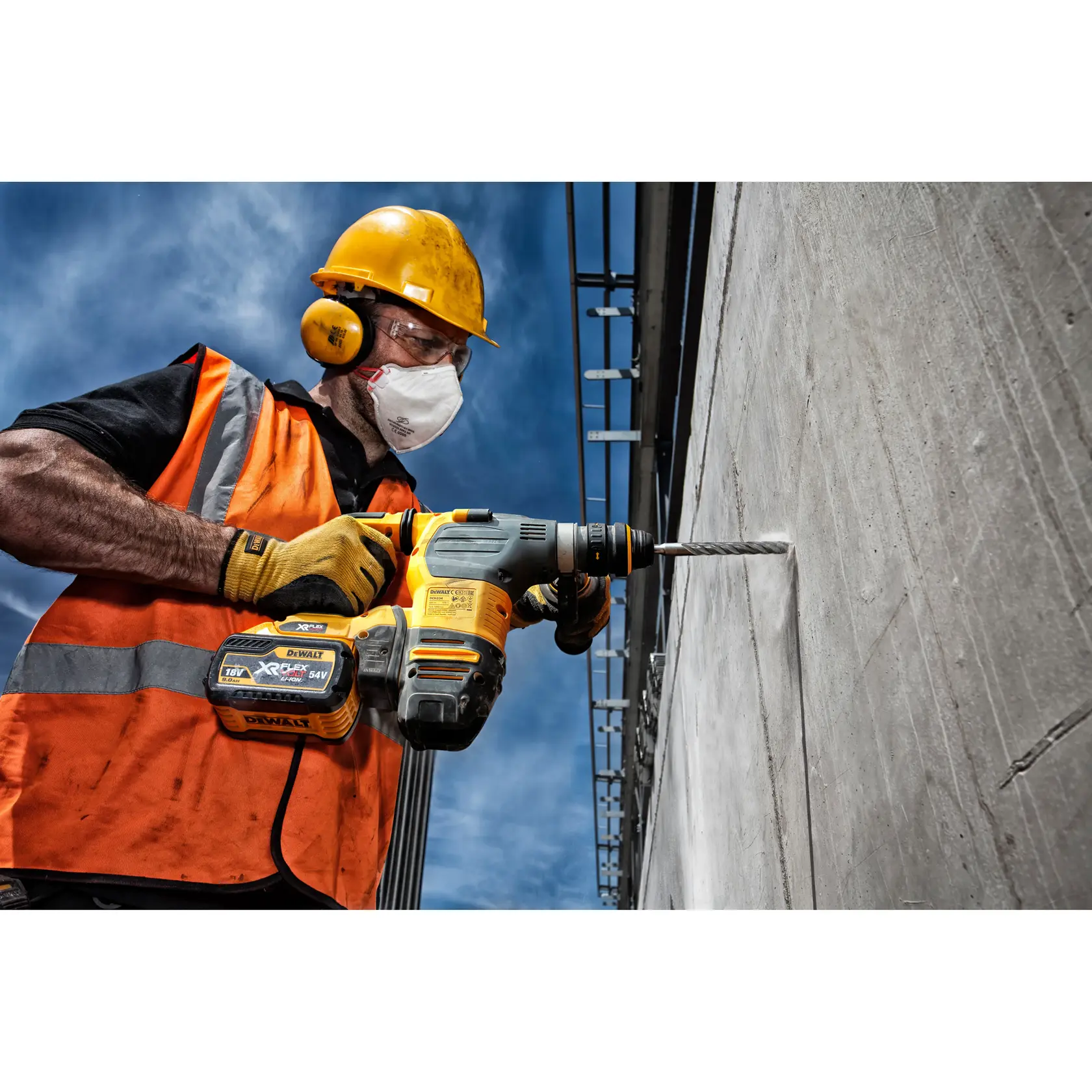 Trabajador perfora muro de hormigón con taladro DEWALT inalámbrico, lleva chaleco naranja y equipo de seguridad.