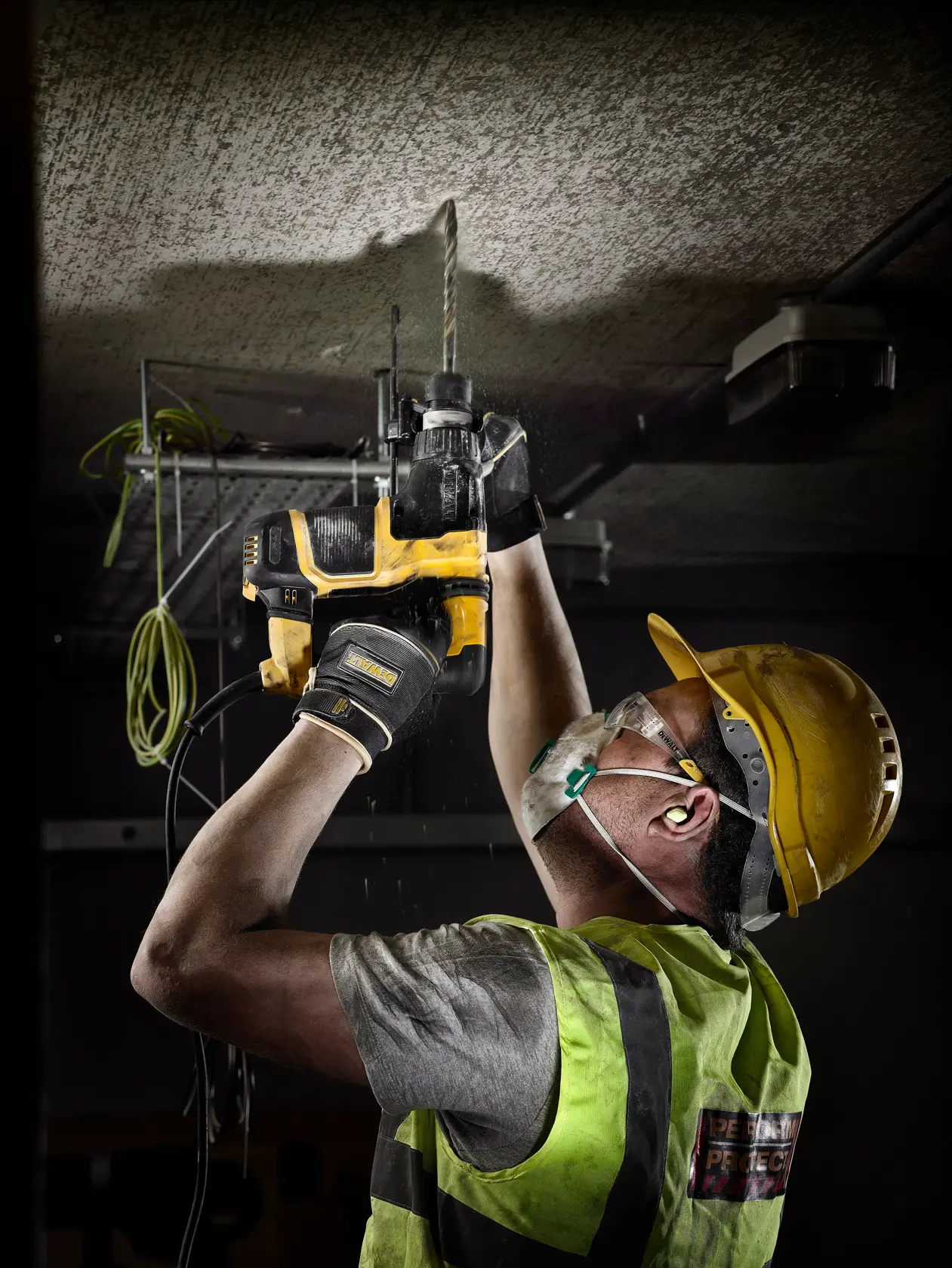 Trabajador perfora el techo con herramienta DEWALT amarilla, lleva equipo de seguridad y chaleco.
