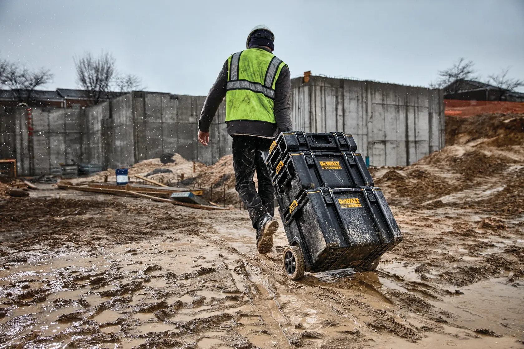 Trabajador arrastra cajas DEWALT con ruedas en obra embarrada, usando chaleco de seguridad y casco.